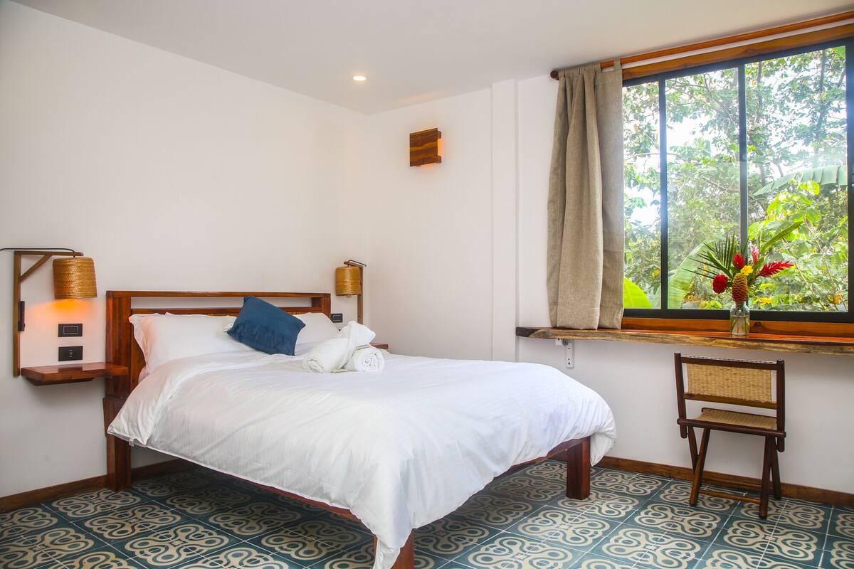 A neatly made bed with white linens, a blue pillow, and a wooden headboard. There is a small table with a lamp and a vase of flowers on the windowsill. The room has patterned floor tiles and a view of greenery outside.