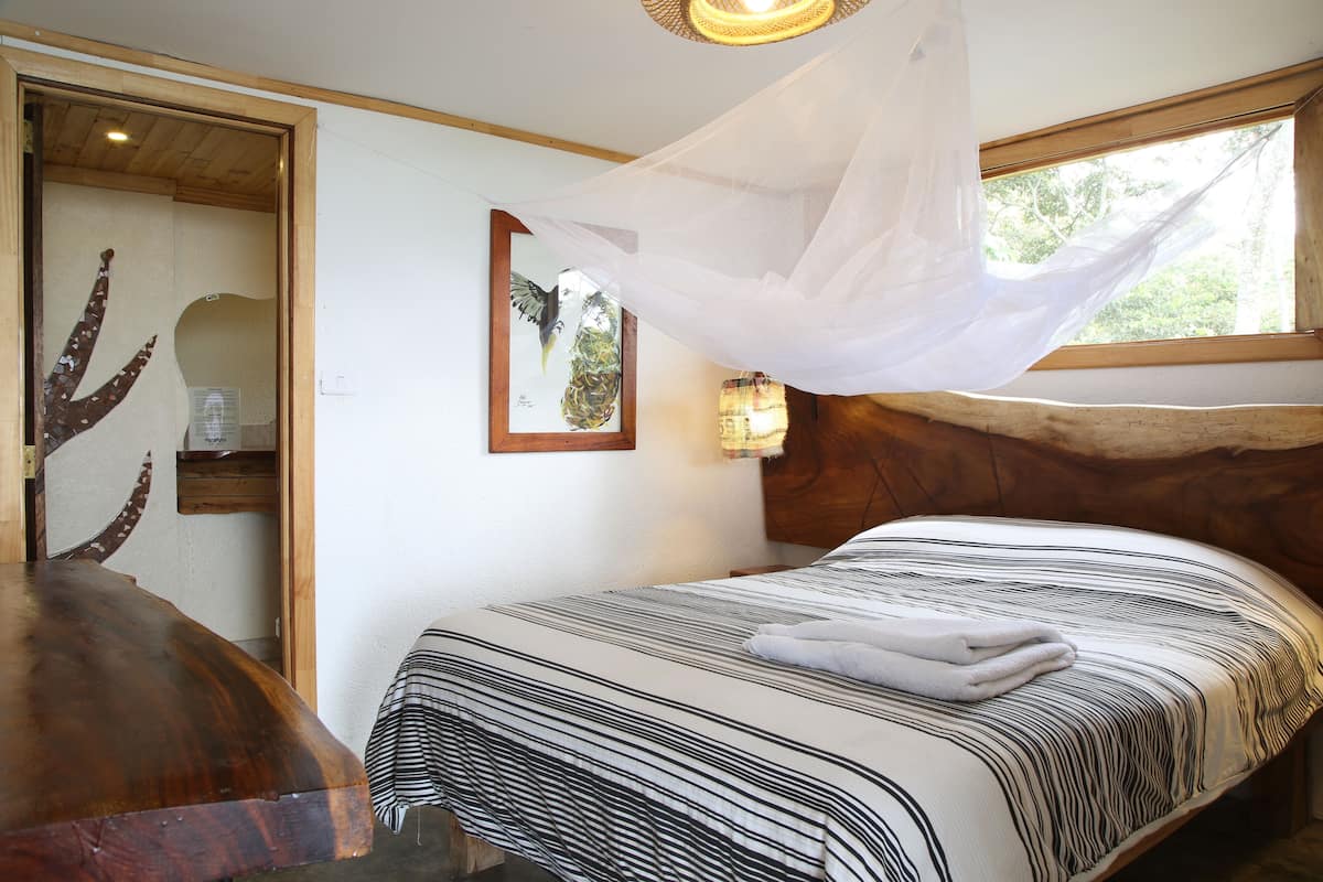 A bedroom with a bed, a wooden headboard, a bedside lamp, a mosquito net, and a framed picture on the wall.
