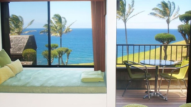 A bedroom with a large window overlooking the ocean, a balcony with a table and chairs, and a wooden floor. - B10: 1 bedroom, 3rd floor, oceanview (Princeville)