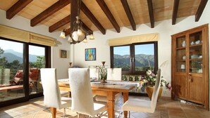 A dining area with a wooden table and chairs, large windows with mountain views, and a wooden cabinet with dishes. - Llobarro, Villa-Finca 5StarsHome Mallorca (Pollensa / Pollença)
