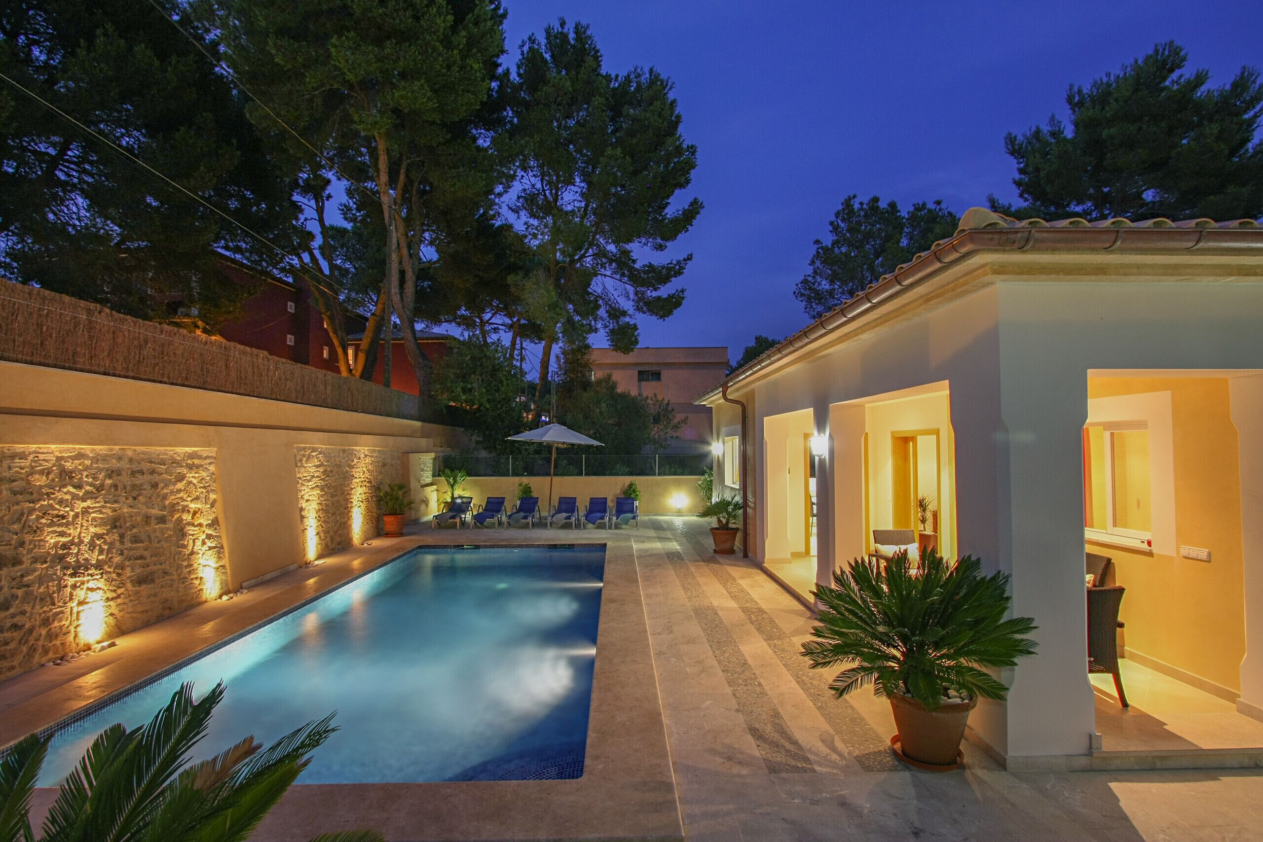 A swimming pool with lounge chairs, a stone wall, and a building with a tiled roof.