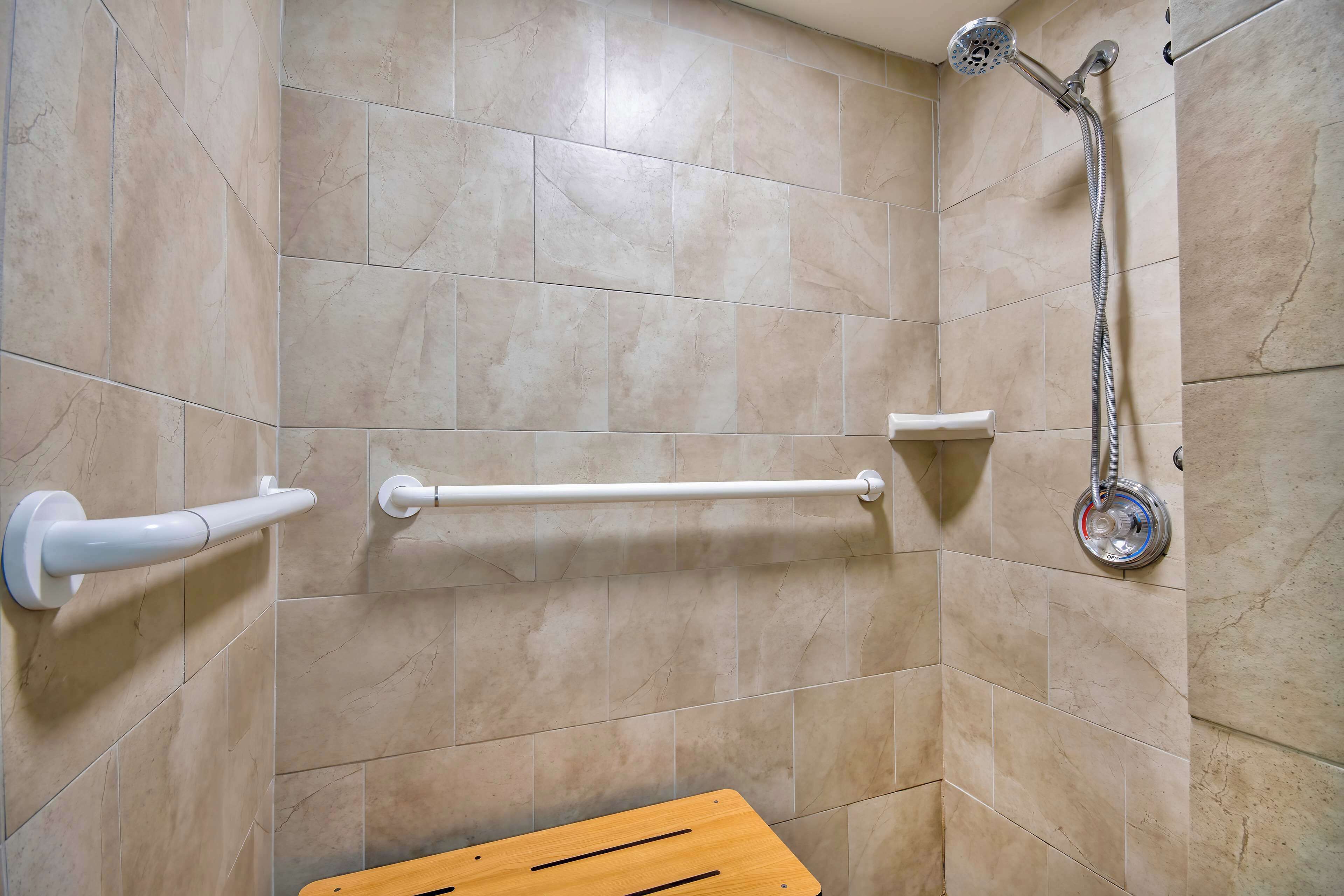 A bathroom with beige tiles, a shower with a handheld showerhead, a white grab bar, and a wooden bench.