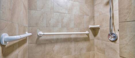 Un baño con azulejos color beige, una ducha con cabezal de ducha manual, un asidero blanco y un banco de madera.