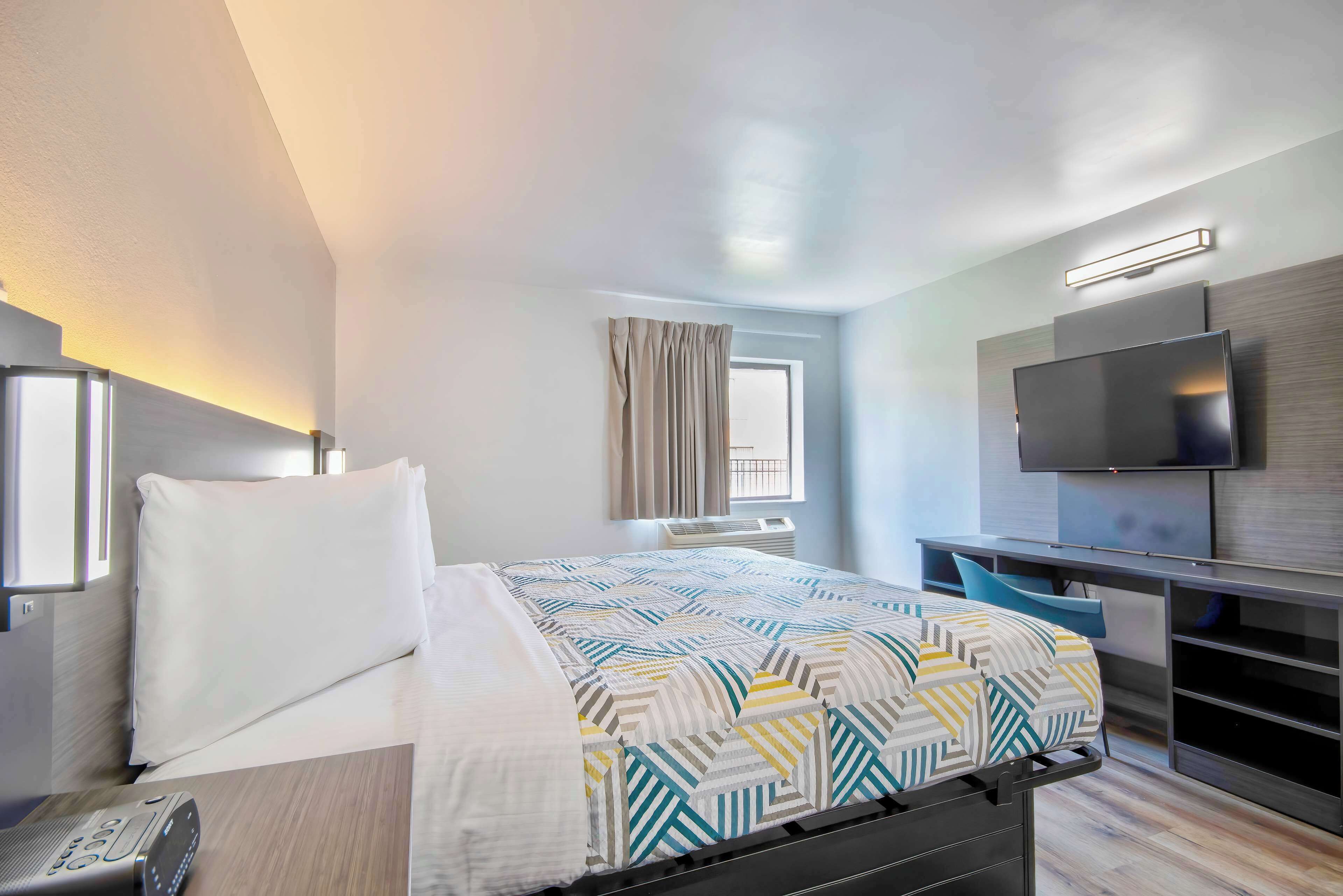 A modern hotel room with a neatly made bed, a desk with a chair, a flat-screen TV mounted on the wall, and a window with curtains.