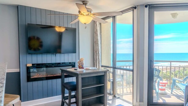 A modern living room with a TV, a ceiling fan, a fireplace, and a balcony with a view of the ocean. - Meridian 705 (Myrtle Beach)