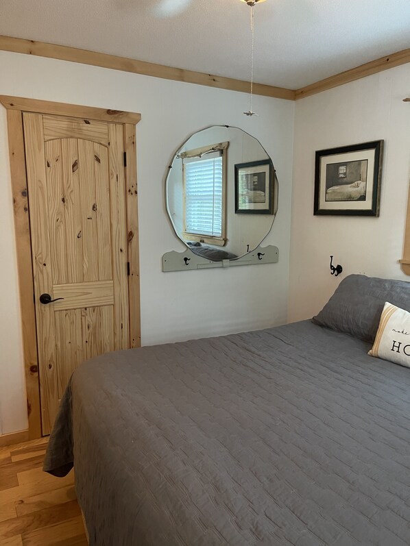 A bedroom with a wooden door, a bed with a grey bedspread, a round mirror, and a framed picture on the wall.