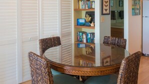 Ein runder Glastisch mit Korbstühlen, ein Bücherregal mit Büchern und Dekorationsobjekten sowie ein weißer Kühlschrank in einem Raum mit Holzböden.