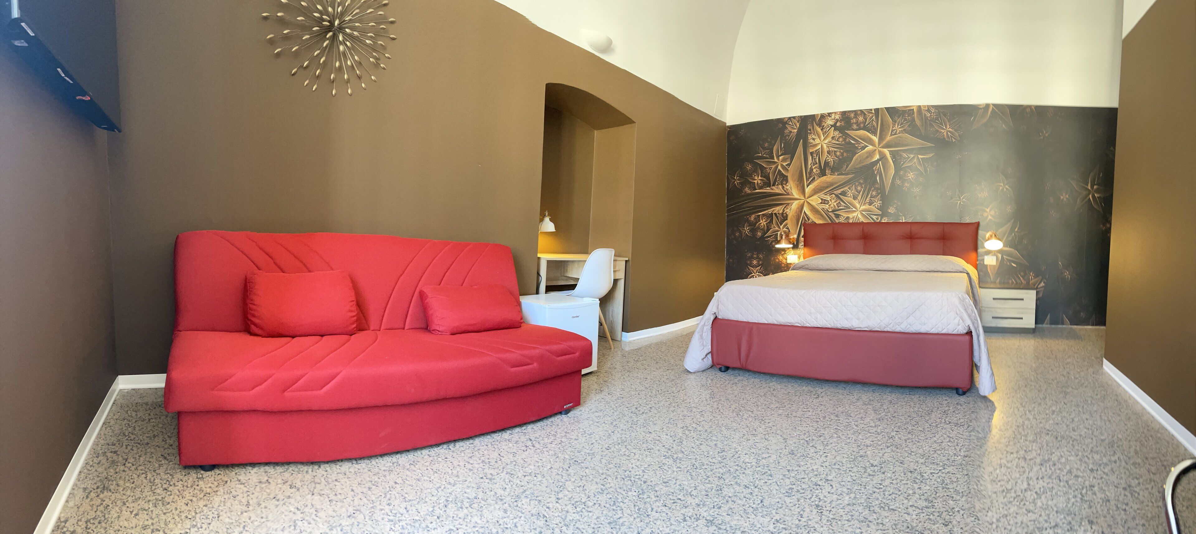 Un dormitorio con un sofá rojo, una cama con cabecera roja, una mesita de noche y una pared con estampado floral.