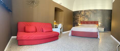 Un dormitorio con un sofá rojo, una cama con cabecera roja, una mesita de noche y una pared con estampado floral.