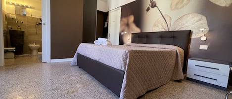 A bedroom with a bed, a nightstand, a wall-mounted light, and a bathroom visible through an open door.