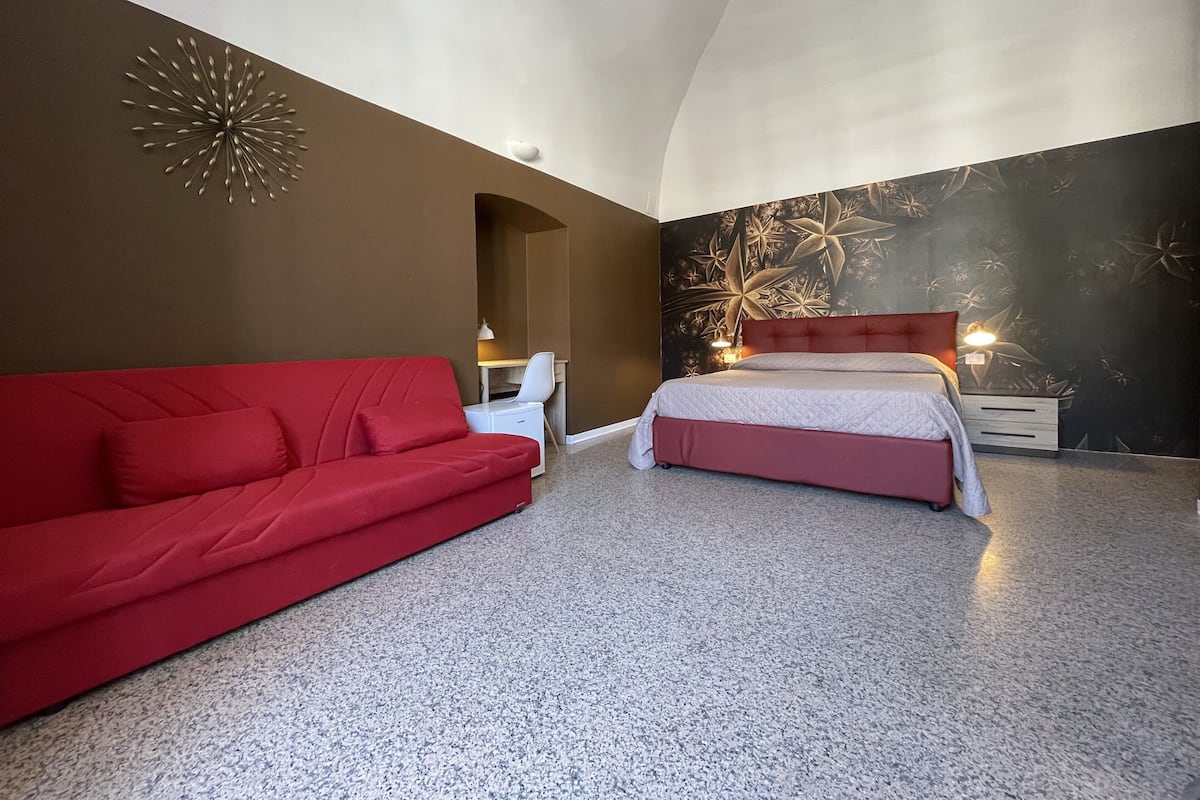 Un dormitorio con un sofá rojo, una cama con cabecera roja, un escritorio y una pared con un diseño decorativo.