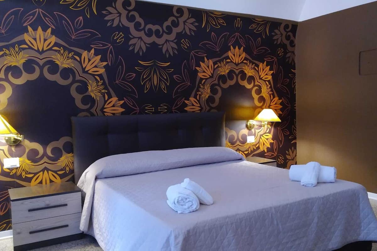 Habitación de hotel con una cama grande, dos lámparas de noche, un cabecero oscuro y una pared decorativa con motivos florales.