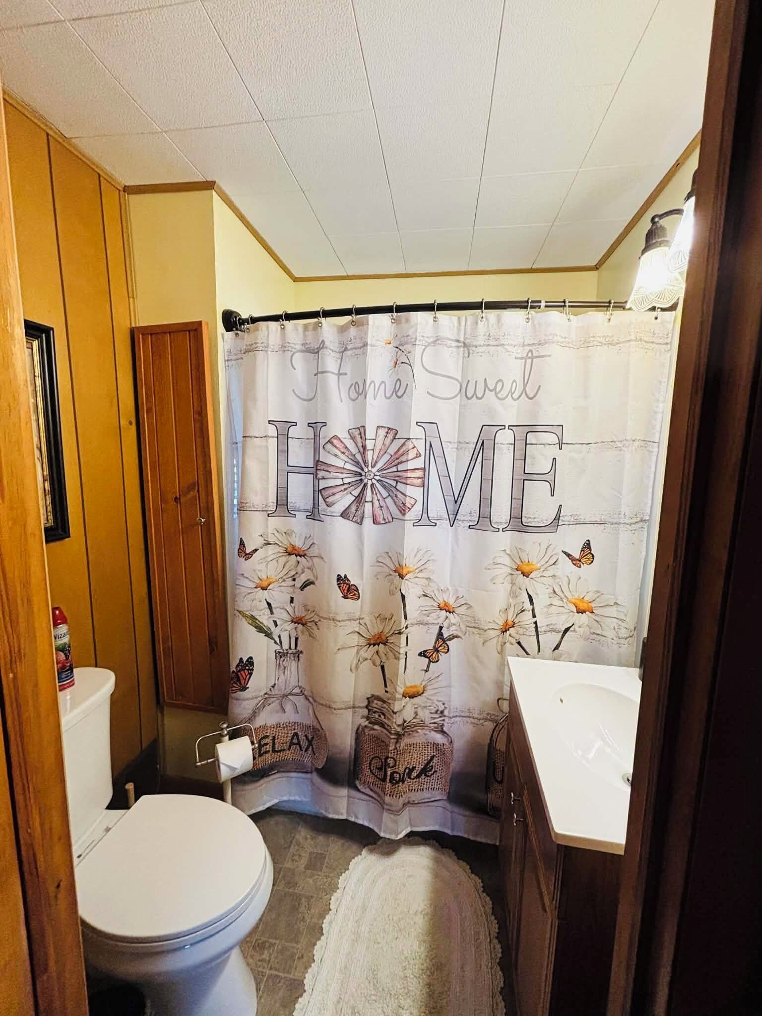Un bagno con tenda da doccia che riporta la scritta 'Home Sweet Home', un water, un lavandino e un tappeto sul pavimento.