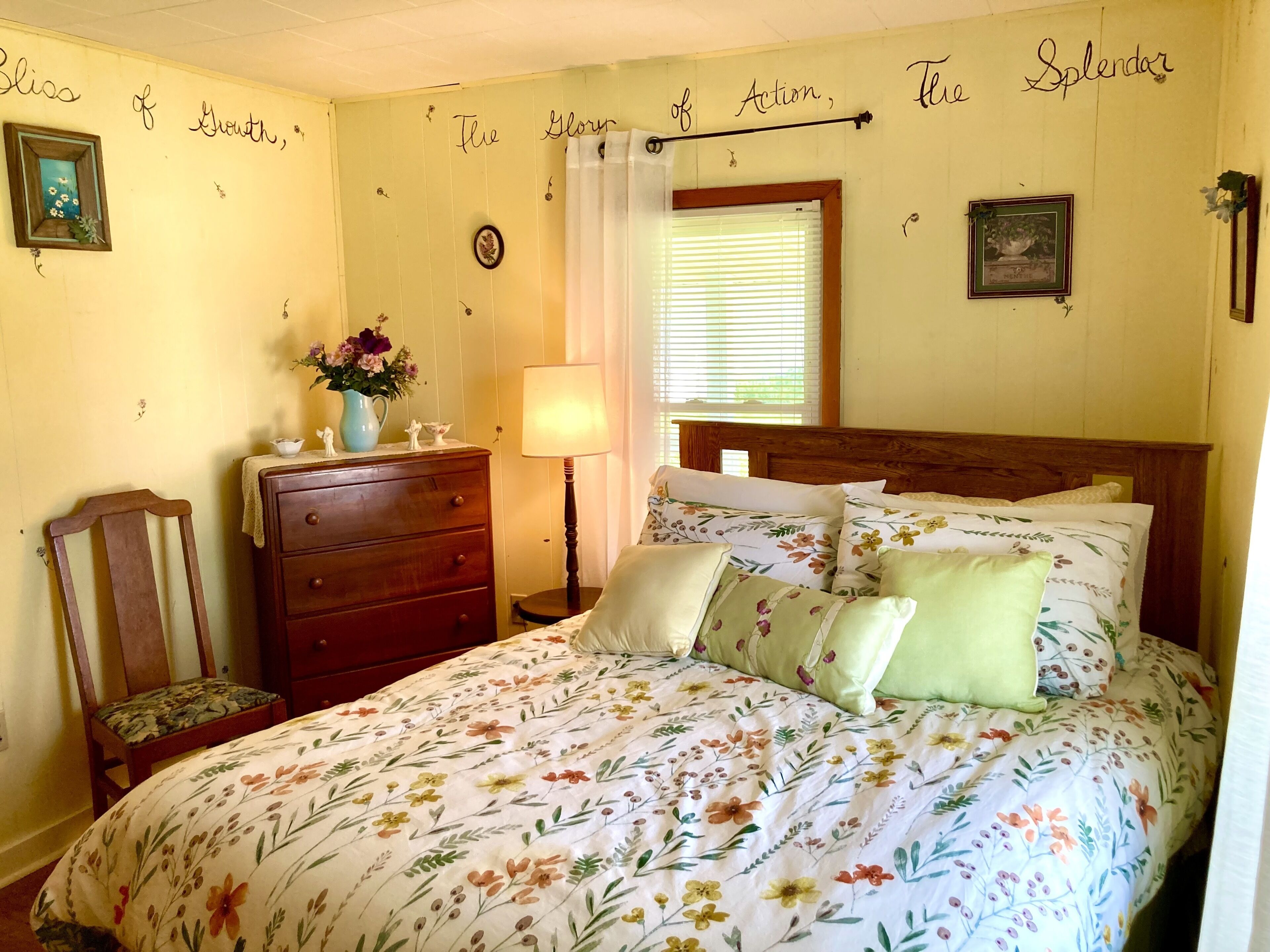 A bedroom with a floral bedspread, a wooden dresser, a chair, and a nightstand with a lamp.