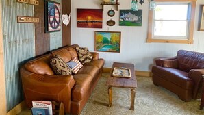 A rustic interior with a brown leather sofa, a wooden coffee table, and wall art including a sunset painting and a house painting.