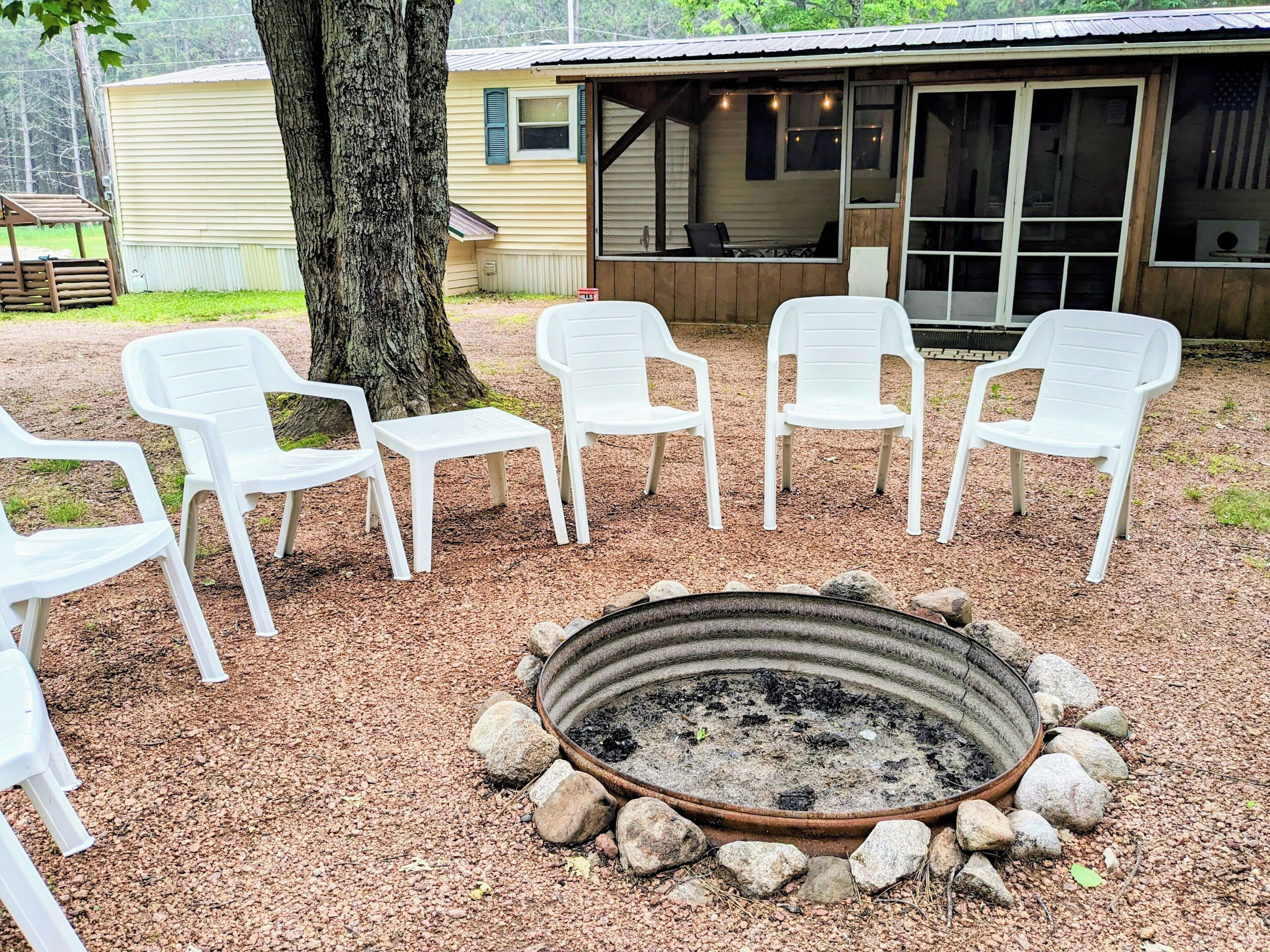 A fire pit surrounded by white plastic chairs and a tree, with a building in the background.