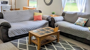 A cozy living room with a grey sofa, a wooden coffee table, and a kitchen area with a microwave. - Backroad Pines Hideaway
Your ATV & Waterfall Adventure Basecamp! (Dunbar)