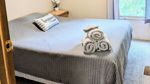 A neatly made bed with a grey blanket and rolled towels, a small bedside table with a vase of flowers, and a window with curtains. - Backroad Pines Hideaway
Your ATV & Waterfall Adventure Basecamp! (Dunbar)