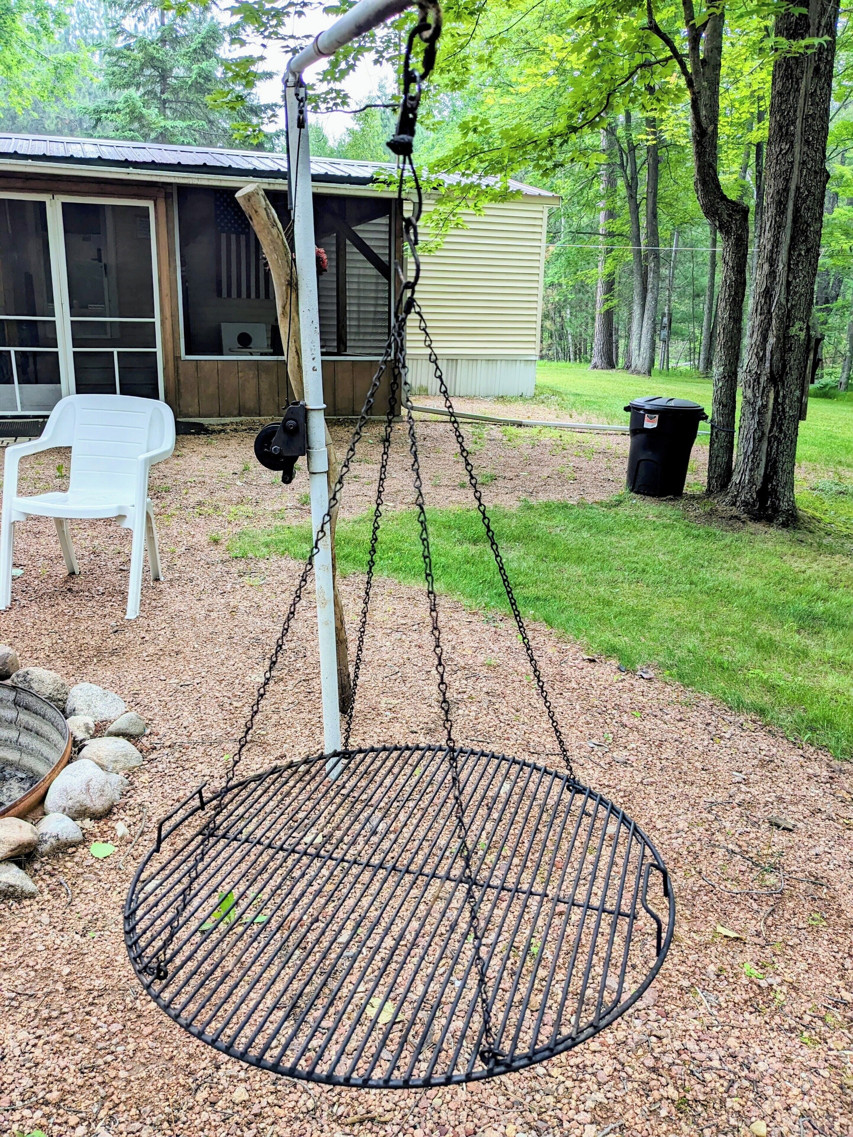 A swing set with a metal frame and chains, a white plastic chair, and a grill in a backyard with a house and trees in the background.