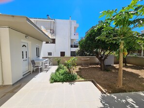 Une cour avec un bâtiment blanc, une table entourée de chaises et un arbre aux feuilles vertes.