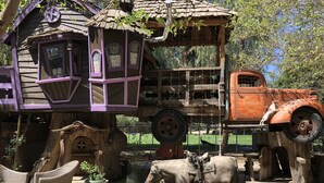 Une construction rustique en bois, dotée d’un toit de chaume, un vieux camion orange et une statue de vache.