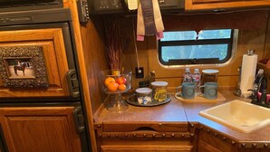 Une cuisine compacte avec des meubles en bois, un évier et une fenêtre. On y trouve un panier de fruits sur le comptoir, ainsi que divers ustensiles de cuisine.