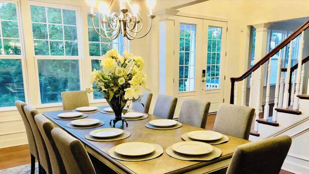 Uma sala de jantar com uma mesa grande posta para seis pessoas, rodeada por cadeiras, um vaso de flores e um candelabro.