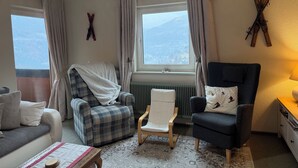 A cozy living room with a plaid armchair, a dark armchair, a white chair, and a sofa. There are two windows with curtains, a radiator, and a wooden floor. - A wonderful panoramic apartment of 110m2. (Radenthein)
