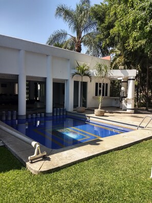 Una piscina con diseño azul y amarillo, rodeada por un edificio blanco con columnas, un porche cubierto y palmeras.