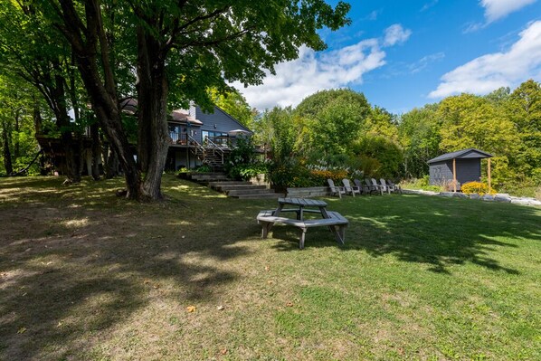 Una casa con terraza, mesa de picnic y un cobertizo pequeño en un área boscosa.