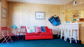 Cozy living space with a sofa, table, and bar stool, featuring a kitchenette and framed artwork.