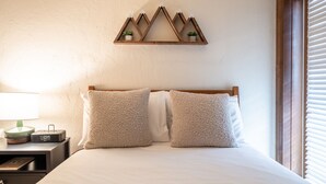 A neatly made bed with two fluffy pillows, a bedside table with a lamp and a digital clock, and a wall-mounted shelf with two small plants. - Cozy Quaint Condo steps away from Vail Village  and  Gondola, Hot tub + Sauna access (Vail)
