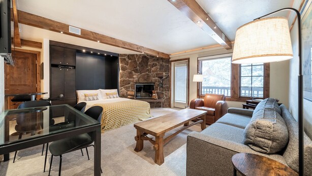 A cozy room with a stone fireplace, a wooden ceiling, a dining table with chairs, a sofa, and a large window with blinds. - Cozy Quaint Condo steps away from Vail Village  and  Gondola, Hot tub + Sauna access (Vail)
