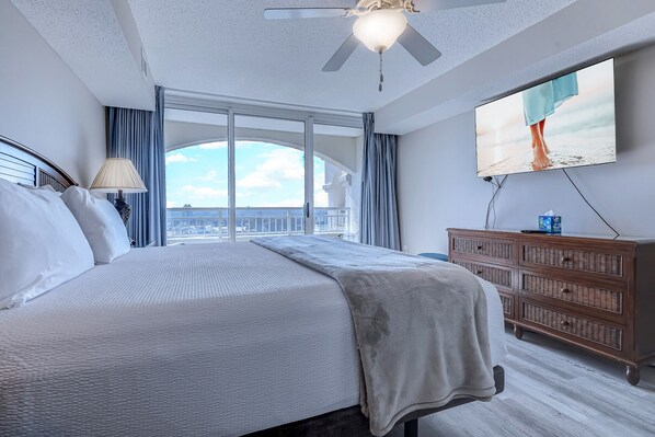 A hotel room with a large bed, a ceiling fan, a TV, and a view of the city through large windows.