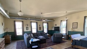 A spacious living room with a large grey patterned rug, a sofa, a chair, and a bed. There are windows with curtains, a ceiling fan, and a television on the wall. - Spacious Loft in Downtown Lewiston  (Lewiston)
