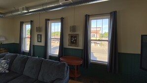 A room with a sofa, a small table, a window with curtains, a wall-mounted air vent, and a framed picture. - Spacious Loft in Downtown Lewiston  (Lewiston)