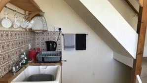 Een kleine keuken met een spoelbak, waterkoker en koffiezetapparaat. Een plank met bekers en een handdoek hangt aan de muur.