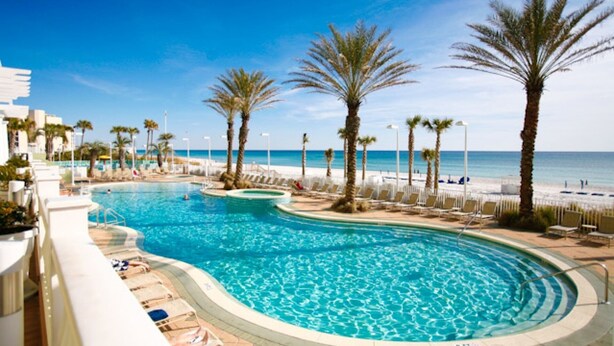 A swimming pool with a curved design, surrounded by lounge chairs and palm trees, with a beach and ocean in the background. - Boardwalk Beach Resort Unit 905 (Panama City Beach)
