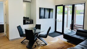 A modern living room with a glass dining table, black chairs, a leather sofa, and a large window with a view of greenery. - Private modern luxury vacation rental (Stockton)