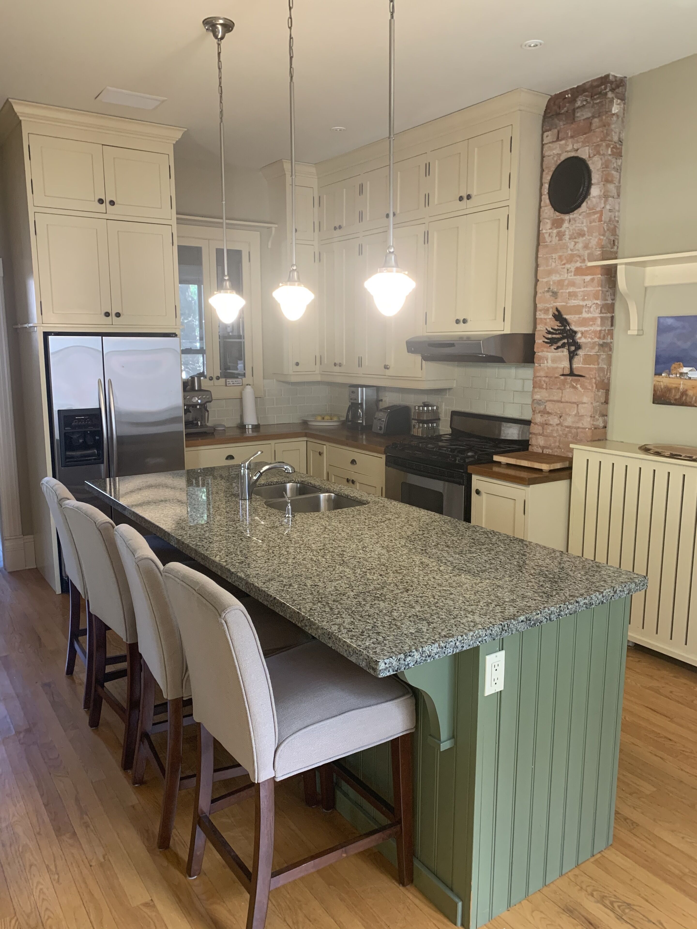 A kitchen with a granite countertop, white cabinets, a stainless steel refrigerator, and a brick accent wall.