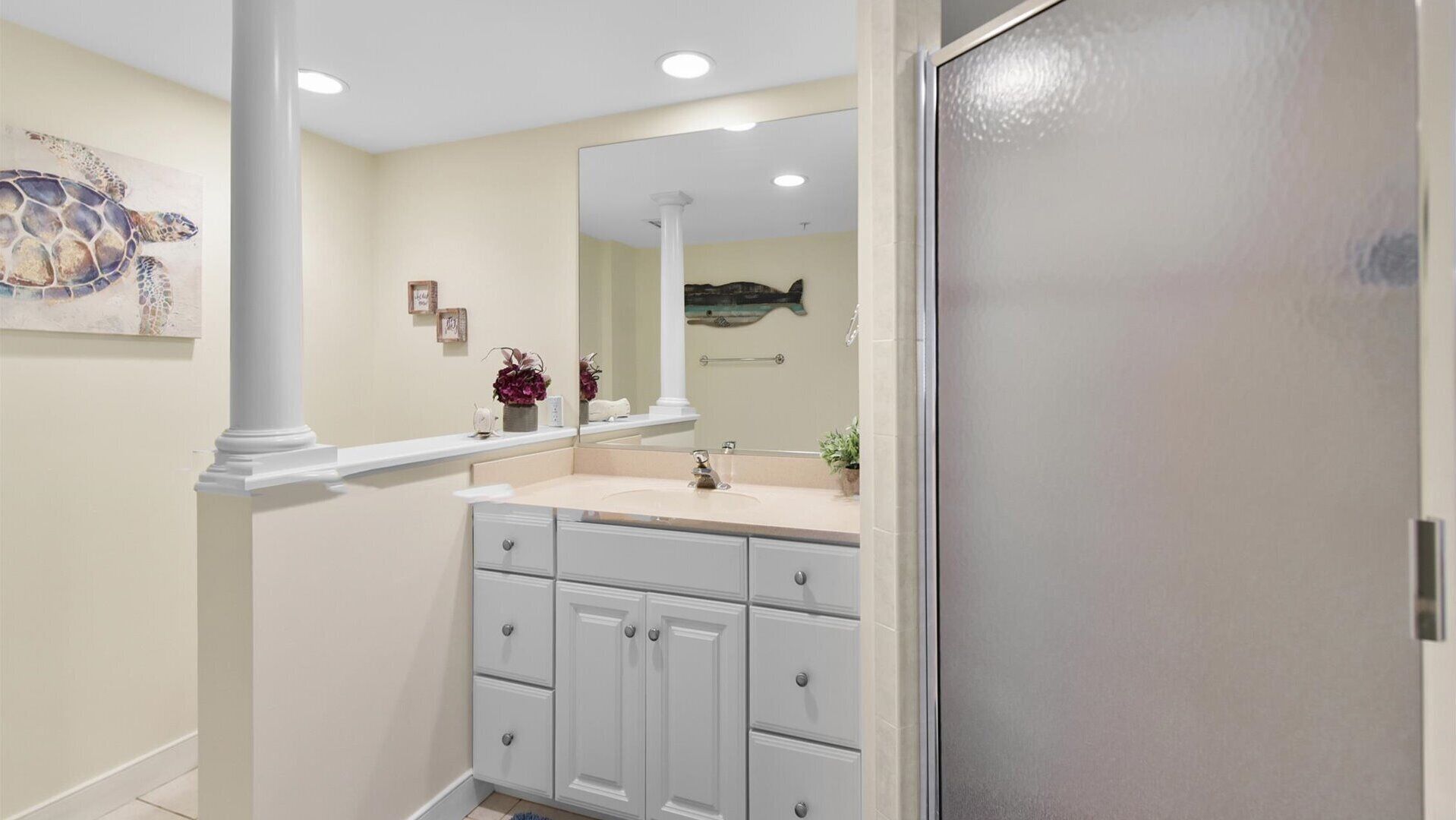 A bathroom with a large mirror, a sink, and a vanity with drawers. There is a decorative piece on the wall and a plant on the counter.