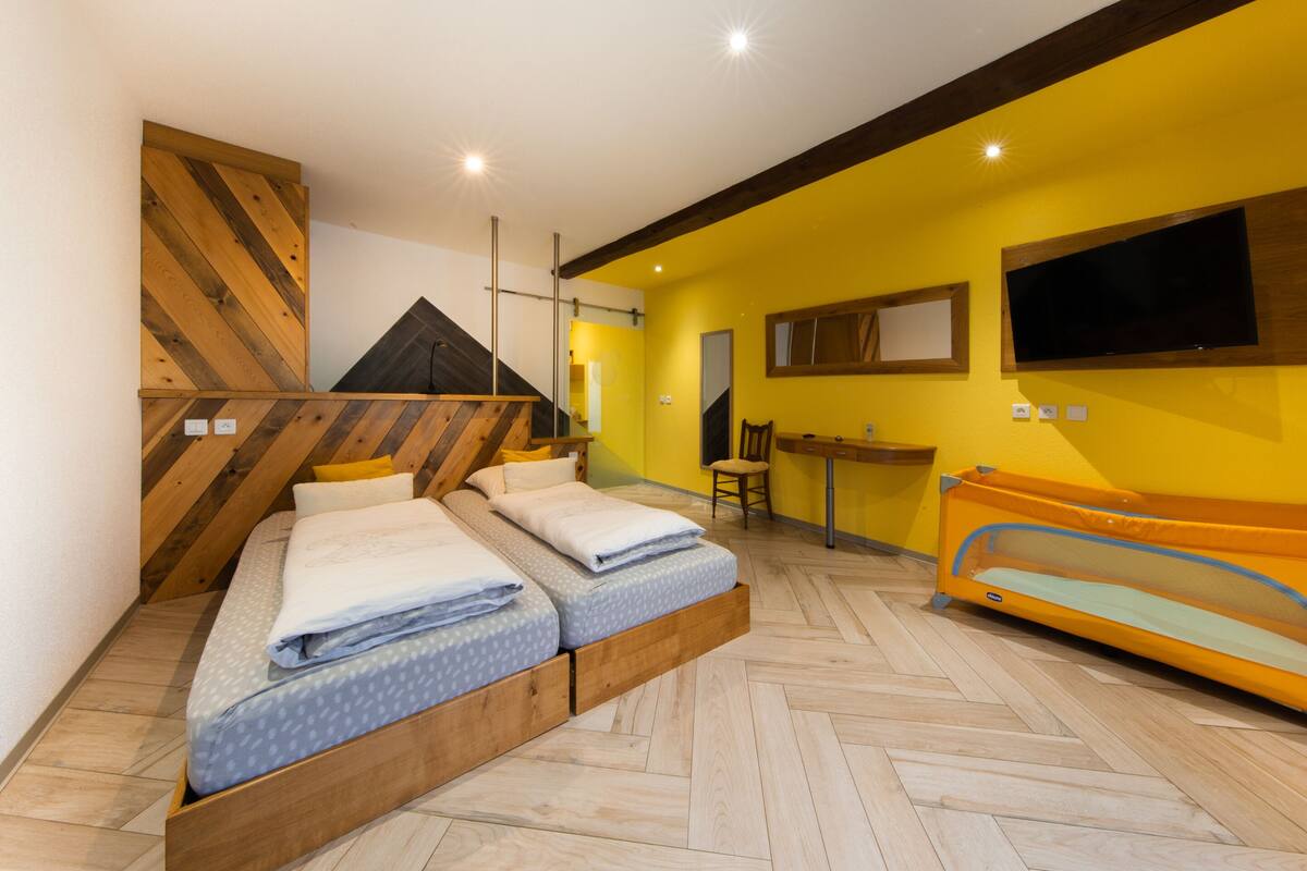 Une chambre à coucher avec un lit, une tête de lit en bois, un bureau, une chaise et un petit parc pour enfants de couleur orange.