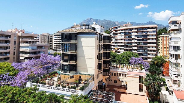 Un edificio residencial de varios pisos con balcones, rodeado de otros edificios y montañas al fondo.