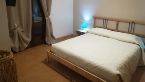 Ein Schlafzimmer mit einem Bettgestell aus Holz, einem Nachttisch mit Lampe und einem teilweise geöffneten Vorhang.