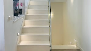 Una escalera con barandilla de vidrio, paredes blancas y un pequeño ventanal en la parte superior.