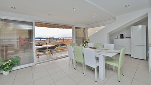 Un área de comedor moderna con una mesa y sillas blancas, vistas a un balcón exterior a través de un vidrio, y una cocina con gabinetes blancos.