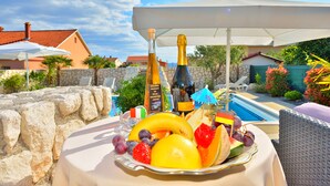 Una mesa con una bandeja de frutas, botellas de champán y un ambiente junto a la piscina con sombrillas y sillones reclinables.