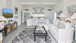 Una sala moderna con un sofá blanco, una mesa de centro y un televisor. Se observa una cocina al fondo con armarios blancos y un comedor.