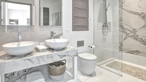 Een moderne badkamer met twee grote witte wastafels, een toilet, een douche met glazen wand en een marmeren spatwand.
