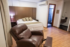 A hotel room with a bed, a leather armchair, a bedside table, and a small desk. - San Diego Suítes Taubaté (Taubate)
